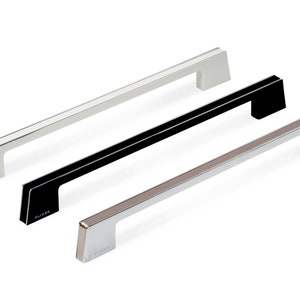 Op de afbeelding: Drie kastdeurgrepen in verschillende afwerkingen: zilver, zwart en wit. Elke greep heeft een rechthoekige vorm met een lichte bocht aan de boven- en onderkant. De grepen zijn allemaal even groot en hebben het woord "Dluker" gegraveerd aan de onderkant.