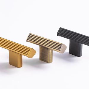 May include: Three cabinet knobs in different finishes: gold, brass, and black. Each knob has a rectangular shape with a grooved design and a T-shaped base.