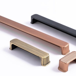 Può includere: Quattro maniglie per armadi in diverse finiture: rame, ottone, nero e rame. Le maniglie hanno un design a coste e una forma rettangolare.