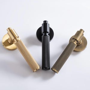 Könnte beinhalten: Drei Türgriffe in Messing, Schwarz und Bronze. Jeder Griff hat einen strukturierten Griff und eine kreisförmige Basis. Die Griffe sind vor einem weißen Hintergrund angeordnet, um ihr Design und ihre Ausführung zu präsentieren. Die Griffe sind für Innentüren.