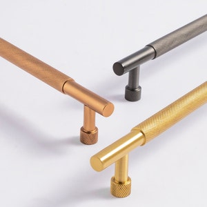 Peut inclure: Gros plan sur trois poignées de meubles aux finitions métalliques différentes : bronze, canon de fusil et or. Chaque poignée a une poignée texturée à motif de losanges et une forme en T. Les poignées sont posées sur un fond blanc.