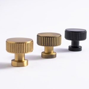 May include: Three cabinet knobs with a ribbed design. The knobs are made of metal and are available in brass and black.