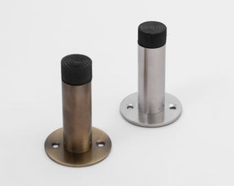May include: Two cylindrical door stops with black rubber tips. One door stop features a brushed bronze finish, while the other has a polished silver-tone metallic finish. Both units include a circular base plate with two pre-drilled screw holes.