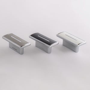 May include: Three rectangular cabinet pulls with a metallic base and a central horizontal groove. The left pull has a brushed silver finish, the center pull is glossy black, and the right pull is polished chrome. Each pull features the text SLIKER.