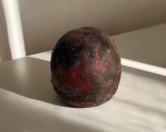 Puede incluir: Un objeto esférico de cerámica con una superficie rugosa y texturizada. El exterior presenta una paleta de colores moteados en tonos rojo óxido profundo, marrón oscuro y negro carbón, creando un aspecto desgastado con una pequeña abertura.