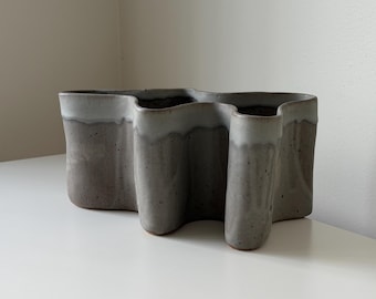Puede incluir: Un jarrón de cerámica gris con un diseño ondulado único. El jarrón tiene una textura moteada y un borde blanco. La forma rectangular tiene tres aberturas, ideal para exhibir flores o como elemento decorativo. Mide unos 30 cm de largo.