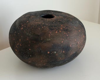 Puede incluir: Una vasija de cerámica redonda y de perfil bajo con una superficie oscura y texturizada que presenta tonos negros y marrones moteados. El exterior está salpicado de pequeños puntos de color naranja vibrante y tiene una abertura circular.