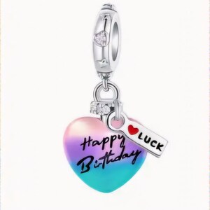 Op de afbeelding: Zilveren bedel met een hartvormige hanger die een kleurverloop van roze, paars en blauw heeft. Het hart heeft de woorden "Happy Birthday" erop geschreven. Een klein label met een rood hart en het woord "LUCK" is aan de bedel bevestigd.