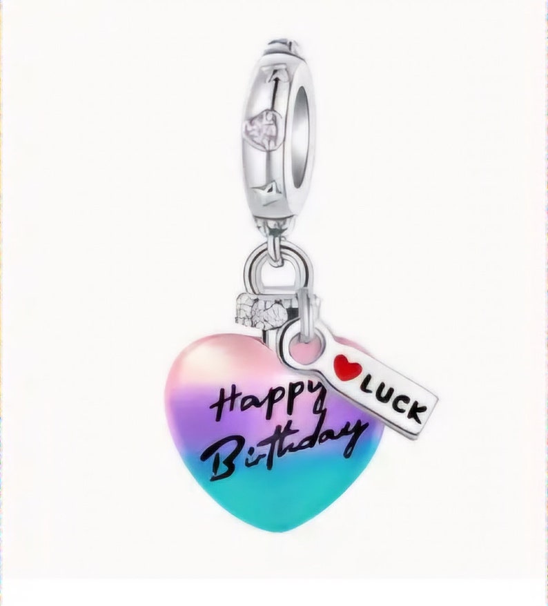 Op de afbeelding: Zilveren bedel met een hartvormige hanger die een kleurverloop van roze, paars en blauw heeft. Het hart heeft de woorden "Happy Birthday" erop geschreven. Een klein label met een rood hart en het woord "LUCK" is aan de bedel bevestigd.