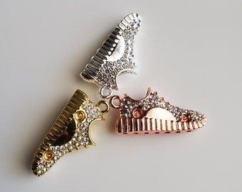 Op de afbeelding: Drie schoenbedels, een gouden, een zilveren en een roségouden, elk met strass.