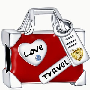 May include: Silver charm in the shape of a red suitcase with a silver handle and a tag that says "Travel". The suitcase has a heart on the front that says "Love" with a blue rhinestone in the center. The tag has a yellow rhinestone in the center.