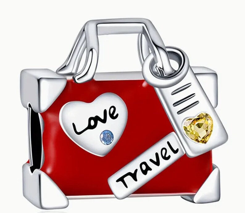 May include: Silver charm in the shape of a red suitcase with a silver handle and a tag that says "Travel". The suitcase has a heart on the front that says "Love" with a blue rhinestone in the center. The tag has a yellow rhinestone in the center.