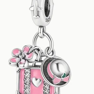 May include: Silver charm with a pink suitcase, a pink flower, and a pink and white hat. The suitcase has a heart and rhinestones. The charm has a silver airplane on the top.