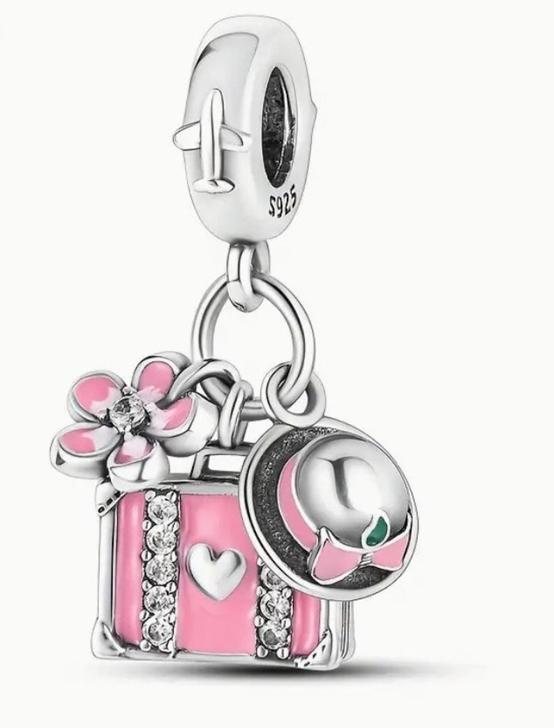May include: Silver charm with a pink suitcase, a pink flower, and a pink and white hat. The suitcase has a heart and rhinestones. The charm has a silver airplane on the top.