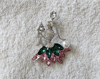 Op de afbeelding: Een zilveren broche met een met kristallen bezette vrouwenfiguur die een parel en een met kristallen bezette 'AKA'-bedel vasthoudt. De jurk van de vrouw is versierd met roze en groene kristallen.