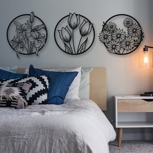 May include: Three black metal wall decor pieces featuring floral designs. The designs include lilies, tulips, and daisies. The decor is hanging on a light gray wall above a white bed with pillows and a nightstand.