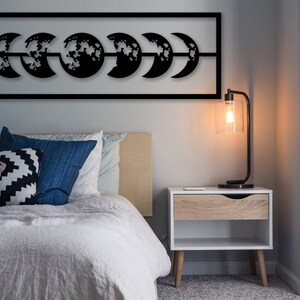 Pode incluir: Uma obra de arte de parede em metal preto retratando as fases da lua. A obra de arte está montada acima de uma cama com almofadas azuis e estampadas. Uma lâmpada moderna com uma sombra transparente está em uma mesa de cabeceira branca.