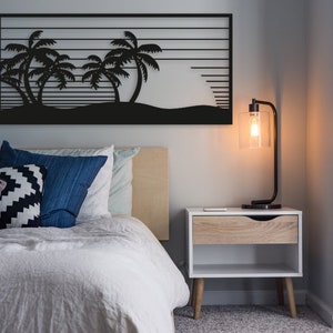 Puede incluir: Arte mural de metal negro con palmeras y un atardecer. La obra de arte está sobre una cama con cojines azules y estampados. Una lámpara moderna se encuentra sobre una mesita de noche blanca.