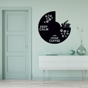 May include: Black wall art with the text "KEEP CALM AND DRINK COFFEE" and coffee-related graphics. The art is a semi-circle shape and is mounted on a light blue wall. A white cabinet and decorative vase are below the art.