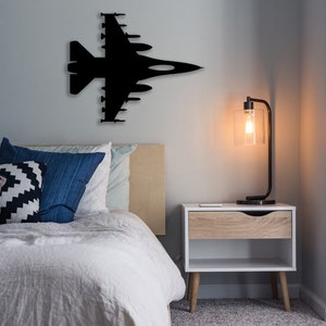 May include: A bedroom with a white bed, black and white patterned pillows, and a black silhouette of a fighter jet on the wall. A wooden nightstand with a lamp sits next to the bed.