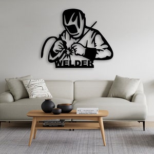 May include: Black metal wall art depicting a welder wearing a welding mask and holding a welding torch. The art is mounted on a white wall above a light grey sofa. The text "WELDER" is below the image.