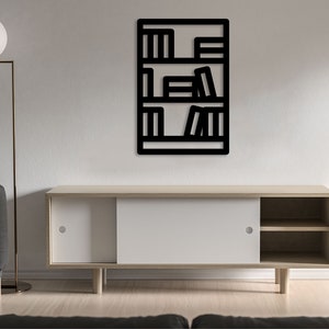 Op de afbeelding: Een zwart metalen wandrek met een minimalistisch ontwerp, met drie planken gevuld met boeken. Het rek is gemonteerd aan een witte muur, met een witte vloer en een grijze bank op de voorgrond.