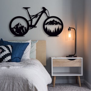Peut inclure: Décoration murale en métal noir représentant un vélo de montagne avec une scène de montagne à l'intérieur de chaque pneu. Le vélo est monté sur un mur blanc au-dessus d'un lit avec des oreillers et une table de chevet.