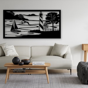 May include: Black metal wall art depicting a lighthouse, sailboat, and trees on a body of water. The art is displayed on a white wall in a living room setting with a couch, coffee table, and ottoman.