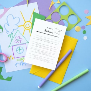 May include: A white sheet of paper with the words "My Name is Bethany" at the top.  The paper has five lines with the word "Bethany" written on each line.  There are two purple and green markers on the paper.  The paper is on a blue surface with a yellow envelope, a drawing of a house, a sun, and a cloud, and a pair of pink scissors.