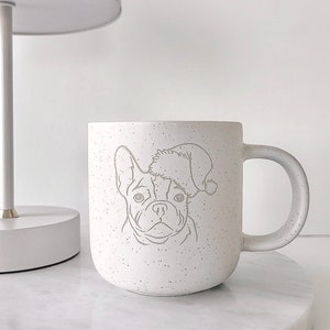 Könnte beinhalten: Eine gesprenkelte weiße Keramik-Tasse mit einer Strichzeichnung einer französischen Bulldogge mit Weihnachtsmütze. Die Tasse hat einen abgerundeten Henkel und steht auf einer weißen Marmoroberfläche. Im Hintergrund ist eine weiße Lampe zu sehen.