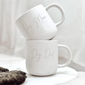 Könnte beinhalten: Zwei weiße Keramikbecher mit gesprenkeltem Überzug. Der obere Becher sagt "Dog Mom" und der untere Becher sagt "Dog Dad".