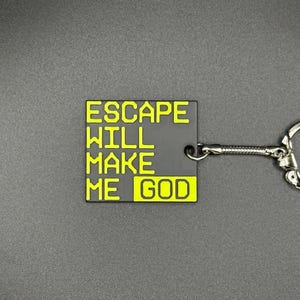 Puede incluir: Un llavero negro con las palabras "ESCAPE WILL MAKE ME GOD" en letras amarillas. El llavero tiene una cadena y un anillo de metal plateado.