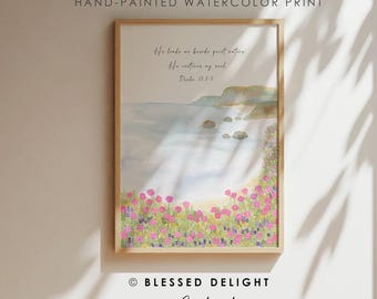 May include: A framed watercolour print featuring a coastal landscape with pink and purple wildflowers in the foreground, a calm blue sea, and the text: He leads me beside quiet waters, he restores my soul. Psalm 23:2-3. The print is labelled Hand-Painted Watercolour Print.
