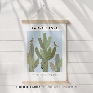 May include: A wall art print featuring a light blue background with a cactus illustration and several birds. The text "FAITHFUL LOVE" is at the top, with a Bible verse below. The print is held by a wooden frame and a string.