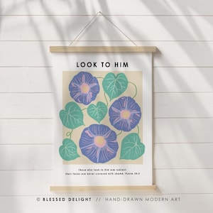 Peut inclure: Une affiche encadrée avec un motif floral. Elle présente des fleurs bleues et roses et des feuilles vertes. Le texte "LOOK TO HIM" est en haut, avec une citation en dessous. L'affiche est suspendue par une barre en bois.