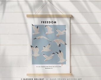 May include: A wall hanging with the word "FREEDOM" at the top. The artwork features white seagulls on a light blue background. The text "It is for freedom that Christ has set us free. Galatians 5:1" is at the bottom. The bottom reads "BLESSED DELIGHT // HAND-DRAWN MODERN ART."