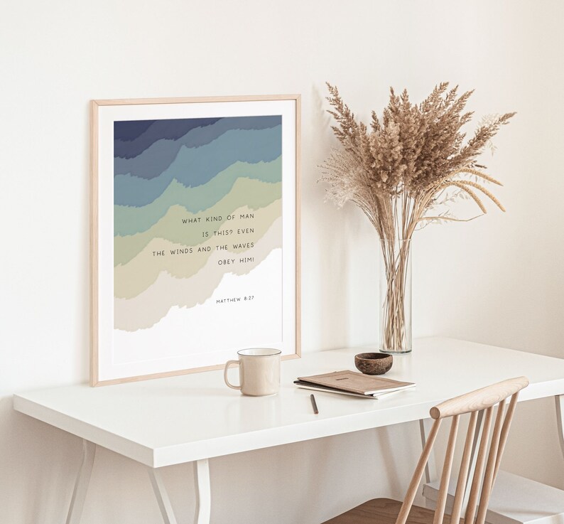 May include: A framed wall art print featuring layered, wavy bands of blue, green, and beige colors. The text reads: WHAT KIND OF MAN IS THIS? EVEN THE WINDS AND THE WAVES OBEY HIM! MATTHEW 8:27. The print is displayed in a light-colored wooden frame.