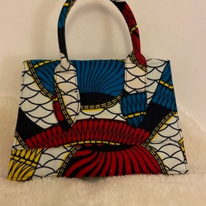 Può includere: Una piccola borsa a mano colorata con un motivo geometrico in tonalità di rosso, blu, giallo e bianco. La borsa ha due manici superiori e una forma strutturata.