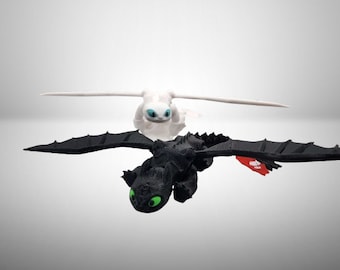 May include: Two dragon figurines, one black and one white, with outstretched wings. The black dragon has green eyes and a red and white emblem on its wing. The white dragon has blue eyes. Both are positioned against a light gray background.