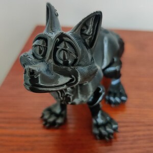 May include: A black 3D-printed figurine of a cartoon-style cat with large eyes and a smiling expression. The cat is standing on all fours. The figurine has a textured surface and is on a wooden surface. The Flexi Factory logo is in the background.
