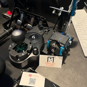 May include: A black and silver racing simulator setup featuring a gear shift, pedals, and a steering wheel mount. The setup includes a black and blue control box with a carbon fiber pattern. A business card with the text "PRA3D Crafts" is also visible.