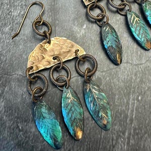 May include: A pair of handcrafted earrings featuring a hammered brass half-moon shape, connected to three dangling teal and bronze leaf-shaped pendants. The earrings have a rustic, bohemian aesthetic, with a dark metal chain.