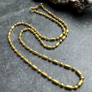 Peut inclure: Un collier doré avec une chaîne composée de petites perles rondes et de perles allongées et cylindriques. Le collier est posé sur une surface gris foncé, avec une formation rocheuse naturelle en arrière-plan.