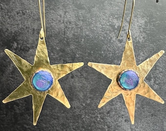 Op de afbeelding: Een paar messingkleurige, stervormige oorbellen met een gehamerde textuur. Elke oorbel heeft een centrale, ronde, iriserende blauw-paarse glazen steen. De oorbellen hangen aan fijne, goudkleurige oorhaakjes.