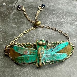 May include: A brass-toned bracelet featuring a dragonfly design with turquoise and bronze accents. The dragonfly is centered on an oval plaque with ornate detailing. The adjustable chain has small links and ends with dark blue beads.