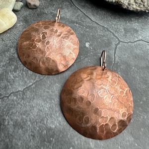 May include: A pair of copper-colored, hammered metal earrings. Each earring is a large, circular disc with a textured surface and a small loop for hanging. The earrings are displayed on a dark gray surface with small rocks in the background.