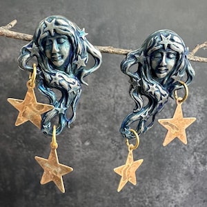 Puede incluir: Un par de pendientes de temática celestial. Cada pendiente presenta una cara en tonos azules con cabello suelto y detalles de estrellas. Un dije de estrella dorada cuelga de cada pendiente, creando un diseño caprichoso y artístico.