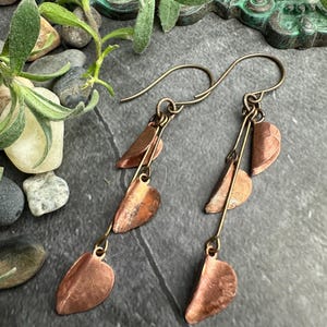 May include: Dangle earrings in copper tones, featuring a leaf design. Each earring has three leaf-shaped elements suspended from a fine wire. The earrings have a rustic, handmade appearance, with a warm, earthy colour scheme.