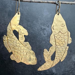 May include: A pair of brass fish earrings. Each earring is a stylized fish shape with etched details, including scales and fins. The earrings hang from simple gold-colored ear wires. The fish are suspended from a thin, brown twig against a dark background.