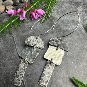 May include: A pair of silver dangle earrings with a hammered texture. Each earring features a half-circle top, a square middle, and a rectangular bottom, all connected by small loops. The earrings hang from curved silver wires. The background includes pink flowers and greenery.